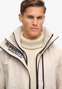 Lys beige jakke med høj krave, lynlås og sort logo. Bæres over en struktureret cremefarvet turtleneck. Skræddersyet design med en glat finish.