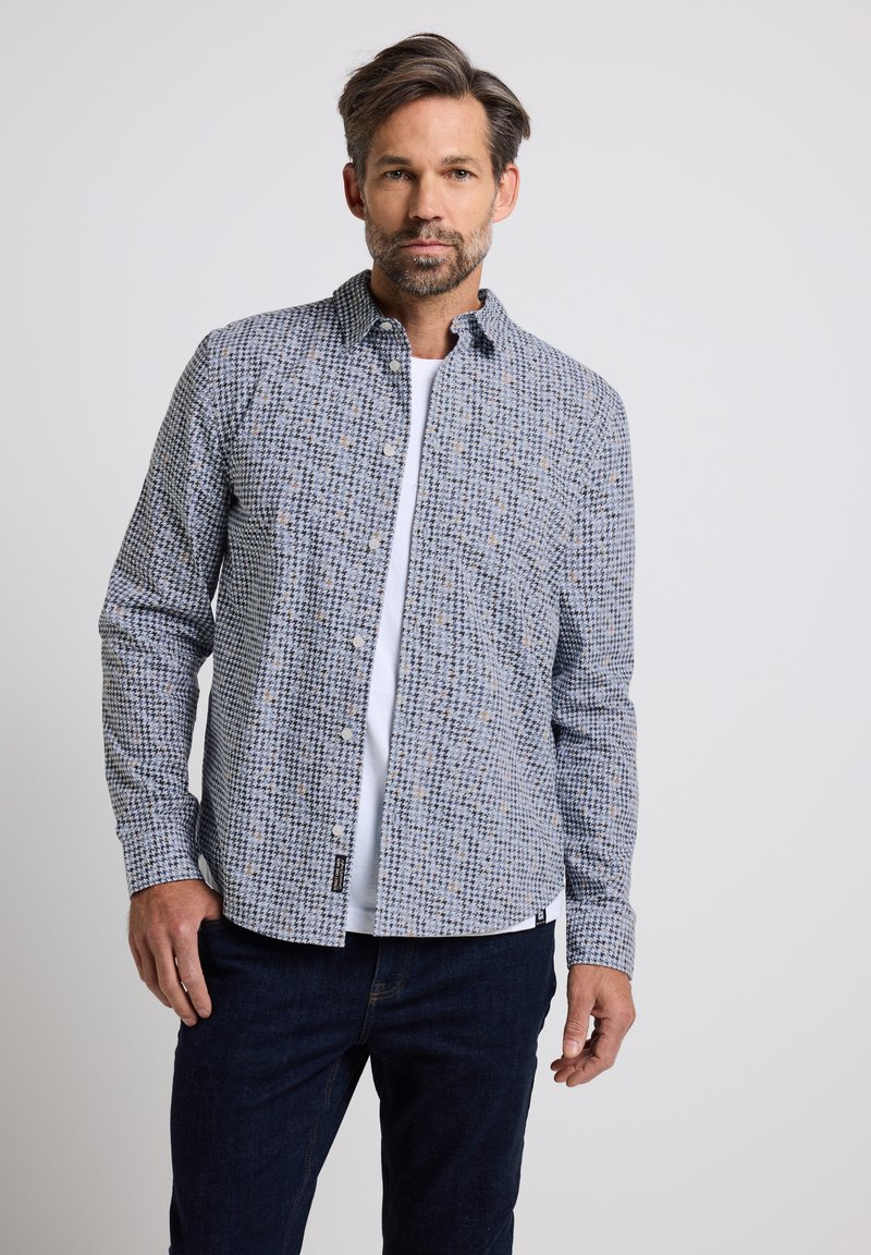 Camicia a maniche lunghe a bottoni blu e bianca con una tasca sul petto, indossata sopra una t-shirt bianca, abbinata a jeans blu.