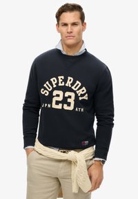 Sudadera marinera con un logo en crema "SUPERDRY 23", cuello redondo, puños acanalados, llevada sobre una camisa de rayas azul y blanca.