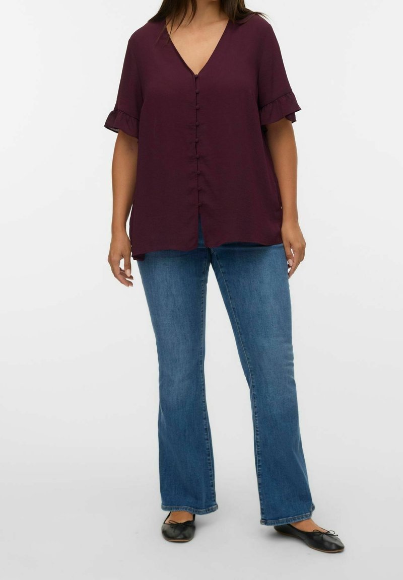 Femme portant un chemisier ample prune à manches courtes avec poignets volantés, jean bootcut bleu et ballerines noires, debout sur fond blanc.