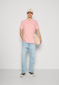 Mladý muž v ružovej polokošeli, svetlomodrých džínsoch, čiernych teniskách, béžovej čiapke, drží béžový klobúk pri bielej stene.