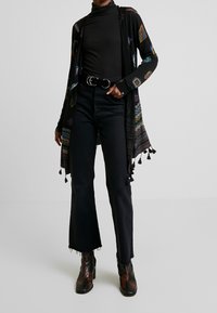 Schwarzer gemusterter Cardigan mit Quastenakzenten über einem schwarzen Rollkragenpullover und hochtaillierten Schlagjeans, ergänzt durch schwarze strukturierte Stiefeletten.