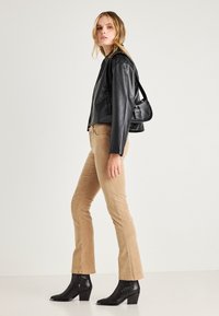 Zwarte leren jas, beige corduroy flared broeken en zwarte enkellaarsjes. Zijprofiel met een kleine zwarte handtas. Eenvoudige texturen en kleuren.