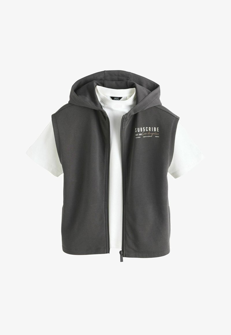 Ciemnoszara bezrękawnik z zamkiem, noszona na białej koszulce z krótkim rękawem, z napisem "SUBSCRIBE Los Angeles" na piersi.