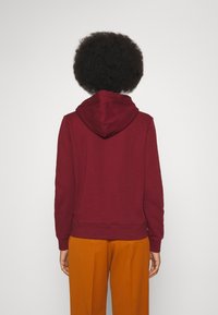 Sudadera burdeos con capucha, bolsillo tipo canguro y puños acanalados, combinada con pantalones anchos naranjas, vista desde atrás.