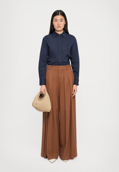 Navy long-sleeve shirt with a pointed collar, paired with high-waisted wide brown pants. Holding a small beige woven handbag.