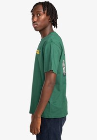 Green cotton t-shirt featuring yellow text and side graphics. Crew neckline, short sleeves, casual fit, and smooth texture.