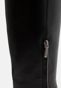 Gros plan d'une botte en cuir noir montrant une fermeture éclair verticale argentée sur le côté avec des détails de couture visibles.