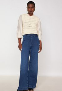 Top in crochet lavorato a maglia di crema con scollatura a onde; abbinato a pantaloni in denim blu scuro a gamba larga a vita alta con cordoncino in vita.
