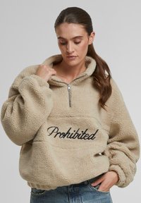 Pullover di pile beige con colletto a metà zip, superficie testurizzata e la scritta "Proibito" ricamata in nero sul davanti. Maniche voluminose.