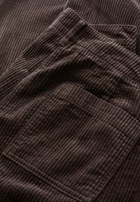 Tmavohnědá manšestrová látka s vertikálním žebrováním, s kapsou a přehnutým pasem, vykazuje jemnou strukturu.