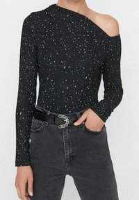 Svart långärmad topp med en axel exponerad, med en ribbad struktur och ett vitt prickigt mönster, tillsammans med högmidjade svarta jeans och ett bälte.