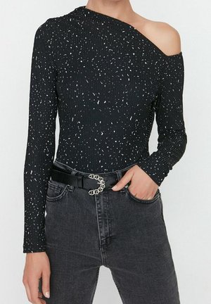 Schwarzes langärmliges Oberteil mit einem freiliegenden Schulter, das eine gerippte Textur und ein weißes Sprenkelmuster aufweist, kombiniert mit hoch geschnittenen schwarzen Jeans und einem Gürtel.