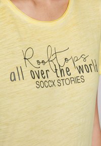 Gelbes T-Shirt mit weichem Material, das schwarzen Drucktext zeigt: „Dächer auf der ganzen Welt“ und „SOCCX GESCHICHTEN“. Rundhalsausschnitt, lässige Passform.