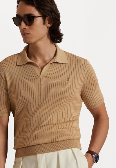 Μπεζ κοντομάνικη πόλο μπλούζα με ριγωτή υφή, με κανονικό γιακά και ένα μικρό κεντητό λογότυπο στο στήθος. Φοριέται με γυαλιά ηλίου.