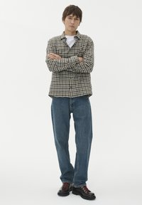 Młody mężczyzna stoi z założonymi rękami, ubrany w brązową koszulę w kratę, białą podkoszulkę, niebieskie dżinsy i ciemne buty z czerwonymi sznurowadłami na białym tle.