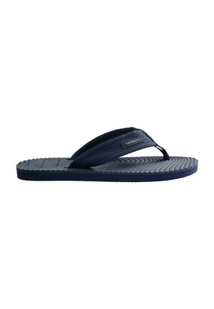 Persoon die lichtroze shorts en hoodie draagt, past een blauwe teenslipper aan terwijl hij op een patroonrijke stenen bestrating bij de zee staat.
