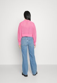 Růžový pletený crop svetr s texturovaným vzorem, žebrované manžety, v kombinaci se světle modrými širokými džíny a černými botami. Pohled zezadu.