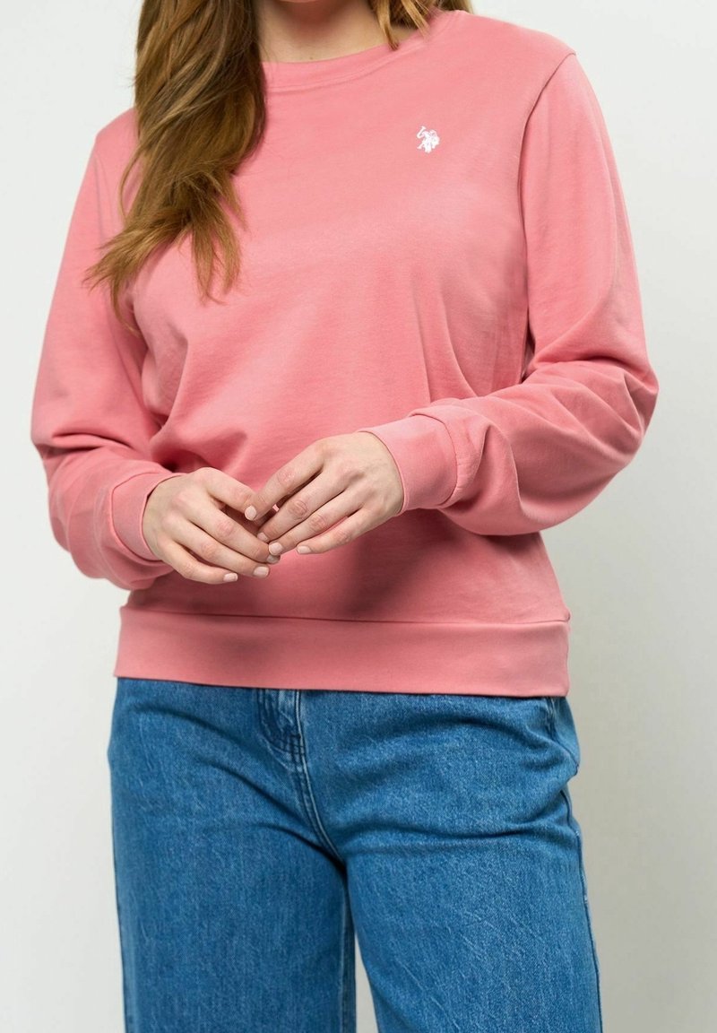 Femme portant un sweat-shirt rose à manches longues avec un petit logo éléphant blanc et un jean bleu, les mains jointes devant.