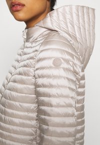 Gros plan sur une personne portant une doudoune à capuche matelassée beige clair avec des coutures horizontales et une fermeture éclair à l'avant, montrant le haut du torse et partiellement le visage.