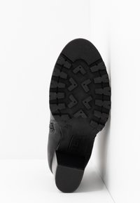 Semelle de botte noire avec des crampons profonds et dessinés ainsi qu'un talon bloc, positionnée verticalement contre un coin blanc en arrière-plan.
