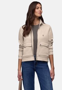 Sweat-shirt beige à fermeture éclair avec deux poches avant, un col montant et un cordon de serrage. Porté par-dessus une chemise grise rayée et associé à un jean bleu.