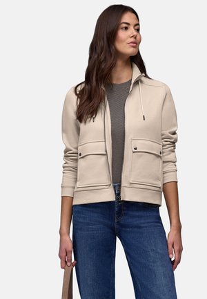 Sweat-shirt beige à fermeture éclair avec deux poches avant, un col montant et un cordon de serrage. Porté par-dessus une chemise grise rayée et associé à un jean bleu.