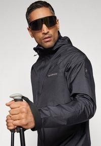 Črna vodoodporna jakna z zadrgo, visokim ovratnikom in detajlom logotipa. Model drži pohodniške palice in nosi velike temne sončne očali. Gladko teksturo.