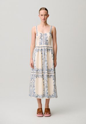 Mujer joven de pie, con un vestido midi color crema con estampados florales azules y sandalias marrones de plataforma con suelas en zigzag rojas.