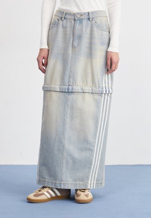 Jupe maxi en denim clair avec bandes blanches latérales, portée avec des baskets beiges et un haut blanc à manches longues, debout sur un sol bleu.