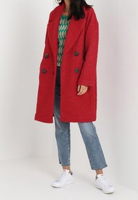 Rød, tekstureret, dobbeltradet frakke med store sorte knapper; båret over en grøn mønstret top, kombineret med blå jeans og hvide sneakers.