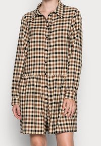 Robe à carreaux avec un devant boutonné, des manches longues et un ourlet à volants. La palette de couleurs comprend du beige, du marron et du noir.