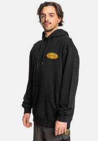 Zwarte hoodie van katoen, met een kangoeroezak en een capuchon met verstelkoord. Gele ovale logo op de borst gedrukt.