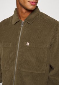 Olivově zelená manšestrová bunda se zipem vpředu, špičatým límcem a dvěma našitými kapsami. Má žebrovanou texturu a malou značkovou cedulku.