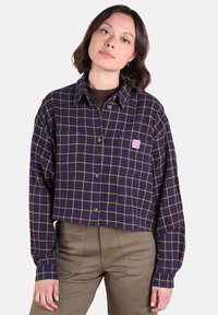 Femme portant un haut court bleu marine avec un motif à carreaux jaunes et un pantalon beige, se tenant devant un fond clair.