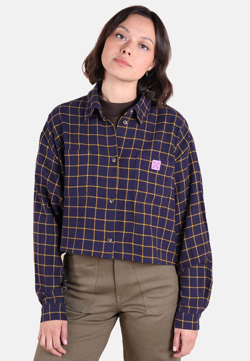 Femme portant un haut court bleu marine avec un motif à carreaux jaunes et un pantalon beige, se tenant devant un fond clair.