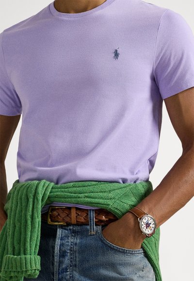 Fialkové tričko s okrúhlym výstrihom z mäkkej bavlny, štýlované zeleným kabelkovým svetrom omotaným okolo pása a zapleteným hnedým koženým opaskom.