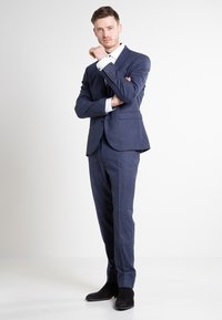 Marineblauer Anzug mit strukturiertem Finish, einreihiger Jacke und passender Hose. Details umfassen ein weißes Hemd und schwarze Wildlederschuhe.
