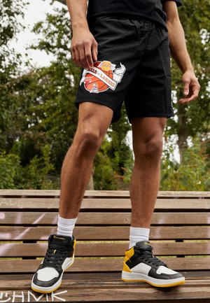 Mann trägt schwarze NBA-Finals-Shorts mit Chicago Bulls vs. Lakers Logo, weiße Socken und schwarze, weiße und gelbe High-Top-Sneaker und steht auf einer Bank.