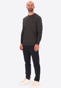 Uomo in piedi, che indossa un maglione grigio scuro a maniche lunghe, pantaloni neri e sneaker bianche, su uno sfondo grigio chiaro uniforme.