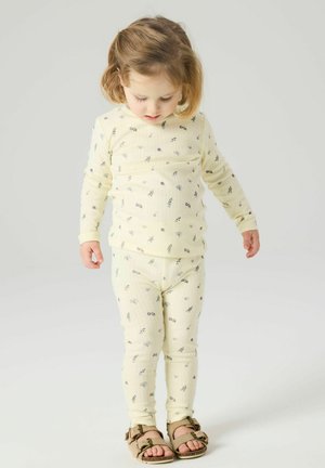 Maluch z jasnobrązowymi włosami, ubrany w kremową kwiatową bluzkę z długim rękawem i spodnie, stoi i patrzy w dół, na nogach ma beżowe sandały.