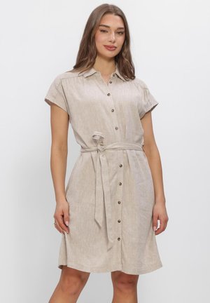 Vrouw draagt een beige jurk met korte mouwen, knopen aan de voorkant en een ceintuur in de taille, staand tegen een effen witte achtergrond.