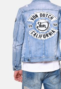 Ljusblå denimjacka med en sliten baksida där "Von Dutch California" är broderad i svart och vitt, knäppta ärmar och en figurnära design.