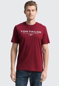 Rode katoenen T-shirt met korte mouwen en witte opdruk met de tekst "TOM TAILOR THE CASUAL DEPARTMENT 1962." Standaard pasvorm, ronde hals.