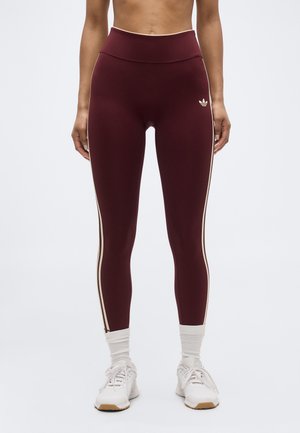 Kvinde iført bordeaux leggings med høj talje og hvide sideløbende striber, hvide crew-sokker og hvide sportssko, stående mod en ensfarvet baggrund.