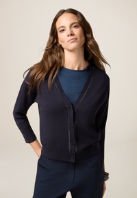 Cardigan blu navy con maniche a tre quarti, fronte texturizzato, chiusura a bottoni e una sottile rifinitura metallica lungo il colletto. Indossato sopra una maglietta blu.
