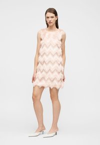Γυναίκα που φοράει λευκά ψηλοτάκουνα παπούτσια και ένα αμάνικο, ανοιχτό ροζ μίνι φόρεμα με υφή και ζιγκ ζαγκ κρόσσια.