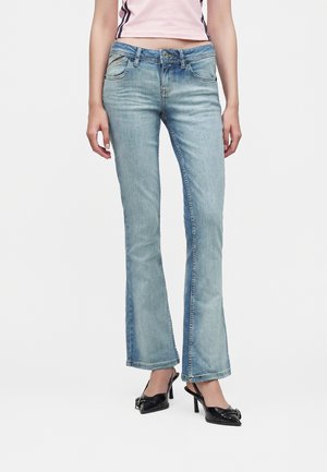 Žena nosi svetlomodré bootcut rifle, ružový krátky top a čierne sandále na opätku so zadným remienkom, stojaca na bielom pozadí.