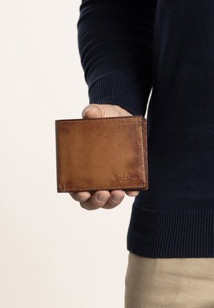 Bruine leren portemonnee met een gladde textuur, bi-fold ontwerp en gestikte randen. Logo is op de voorkant gedrukt. Gehouden in een hand.