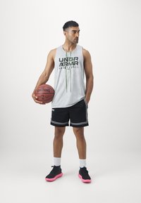 Camiseta de deporte gris sin mangas con el logo negro y verde de "Under Armour Basketball", combinada con pantalones cortos negros con acentos blancos y zapatillas de deporte con suela rosa.